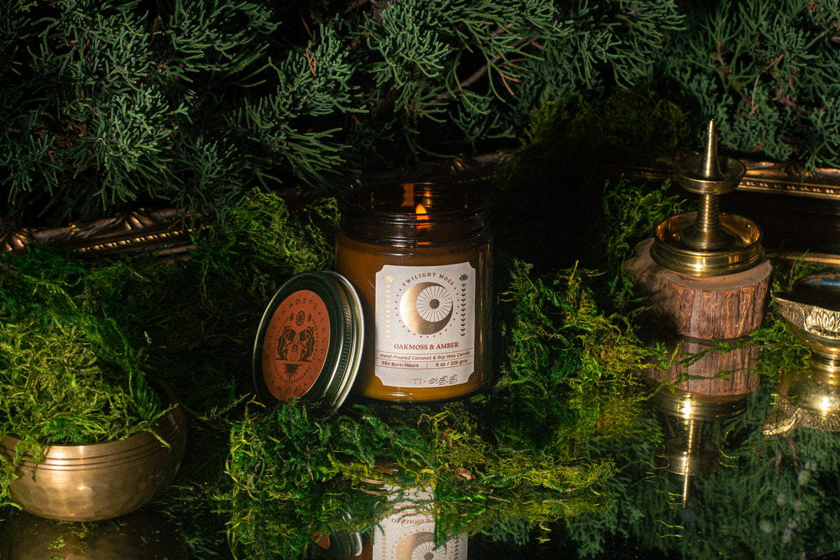 ADITI® Twilight Moss candle among green foliage and brass vessels on a mirrored surface